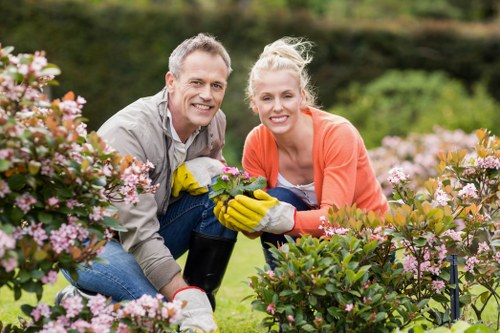 A mature man and woman are kneeling together in a well-maintained garden, surrounded by lush green plants and blooming pink and purple flowers. They are smiling at the camera and appear to be engaged in gardening activities, with the woman wearing yellow gardening gloves and holding a small potted plant, while the man supports her. The garden features neatly arranged flower beds with dense foliage and vibrant blossoms, a background of trees and bushes, and a lightly cloudy sky indicating mild weather. The scene shows an outdoor space designed for relaxation and gardening enjoyment, reflecting the professional landscaping and lawn care services offered by Gardening Services Chessington. The natural tones of greenery and bright floral colours enhance the welcoming and well-kept appearance of the garden, suggesting a high level of horticultural care and attention to detail, typical of local garden maintenance and landscaping projects in the Chessington area.